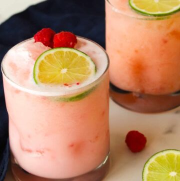 Two rocks glasses of raspberry coconut margarita on a marble counter with lime slices and raspberries.