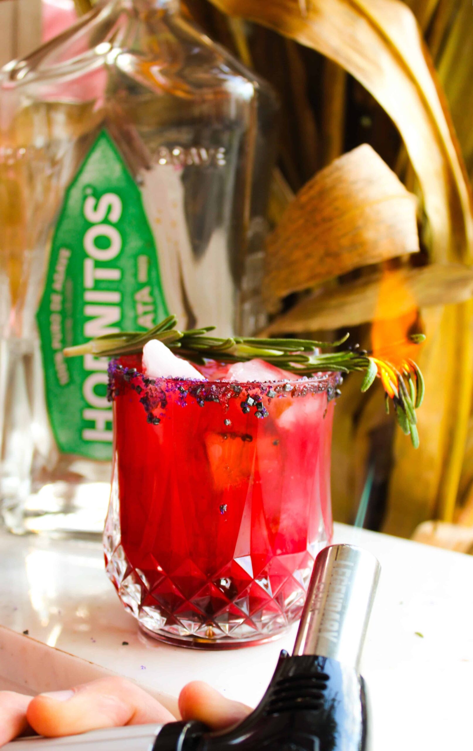 Lighting a rosemary sprig garnish on fire while it rests on top of a red margarita.